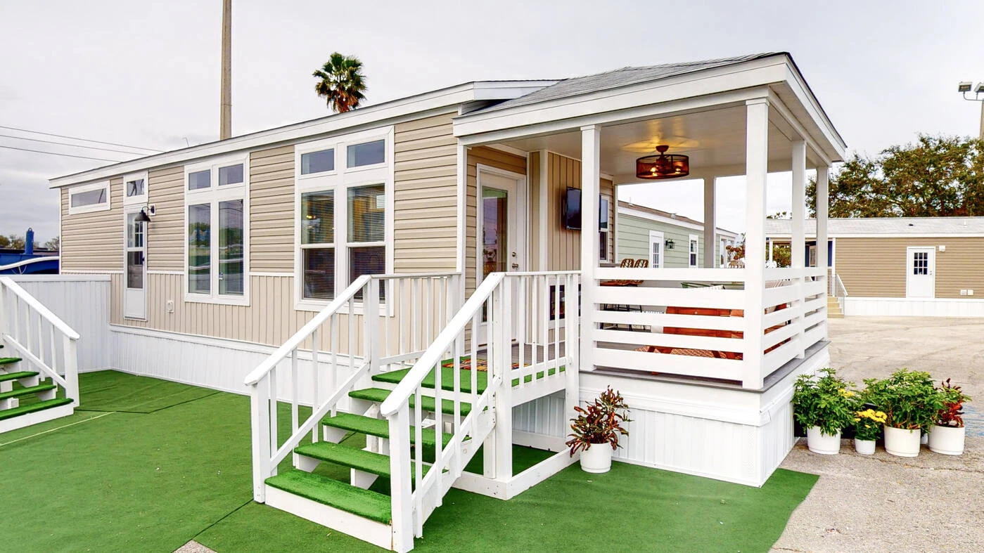 A beige manufactured home with green steps, a covered porch, white railing, and several potted plants on artificial grass.