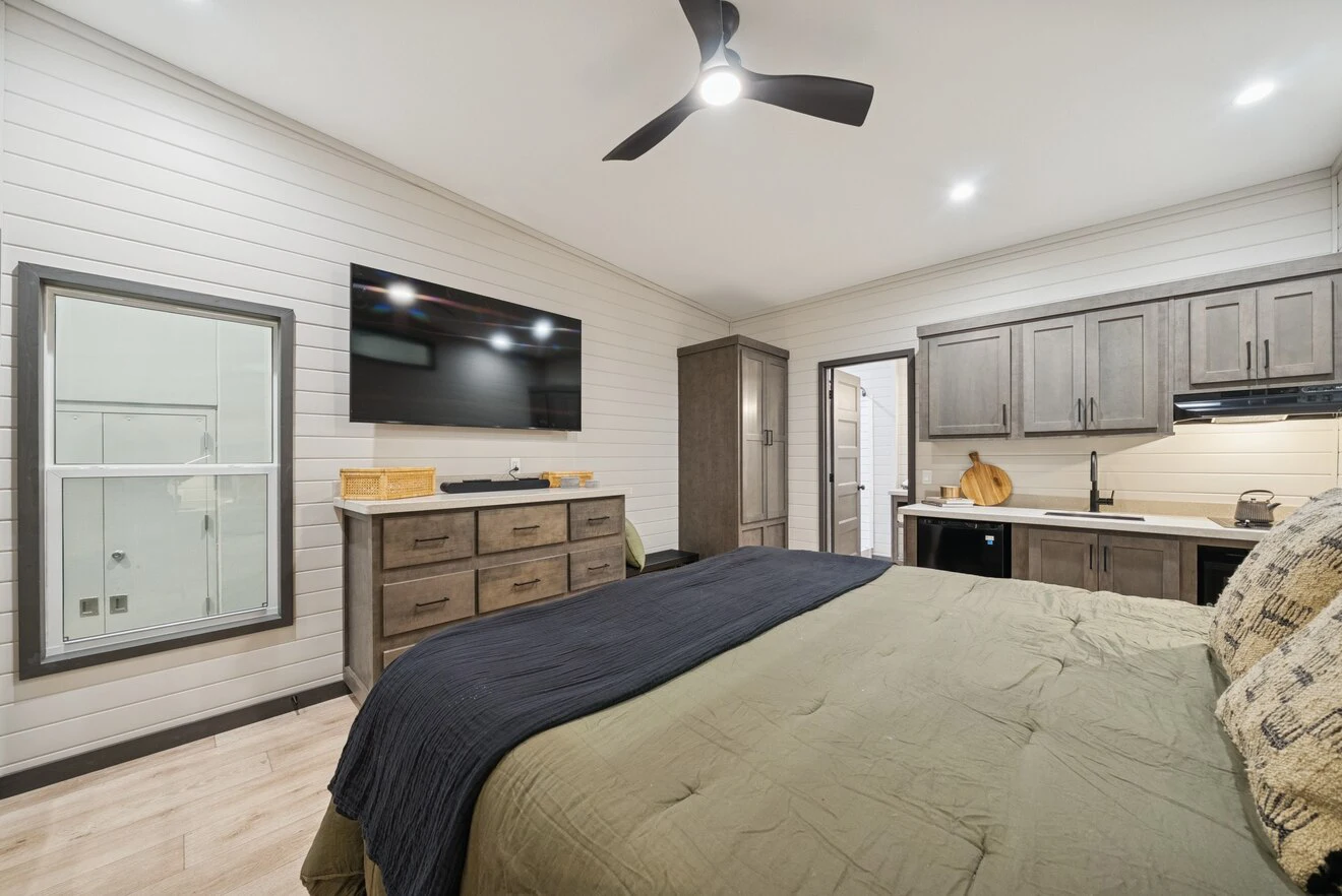 Modern bedroom with a large bed, dresser, wall-mounted TV, kitchenette, and ceiling fan; light wood floors and white shiplap walls create a clean, minimalist look.