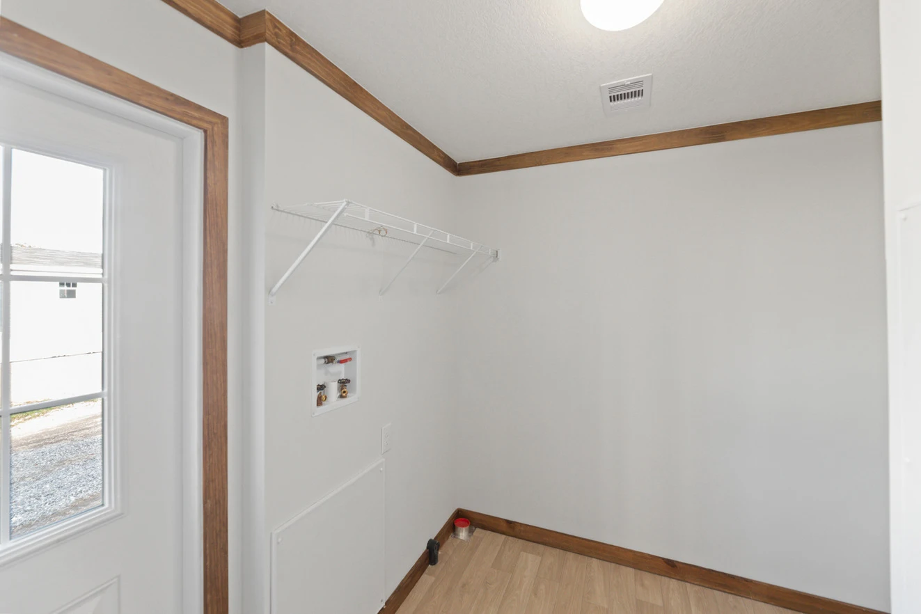 24_ds_290_thegoose_ly28643a_interior_002_web_1738879230305_1323_12 A small laundry room with white walls, wooden trim, a wire shelf, washer and dryer hookups, and a door with a window.