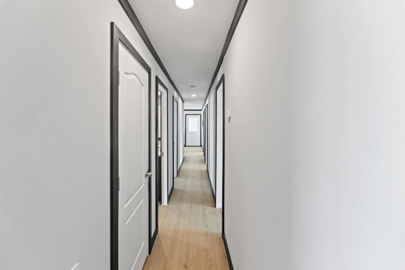 24_ds_290_thesparrow_ly32725a_interior004_web_1738876863711_1323_12 A long, narrow hallway with light-colored walls, wood flooring, and several white doors with black trim on both sides. Ceiling lights illuminate the corridor.