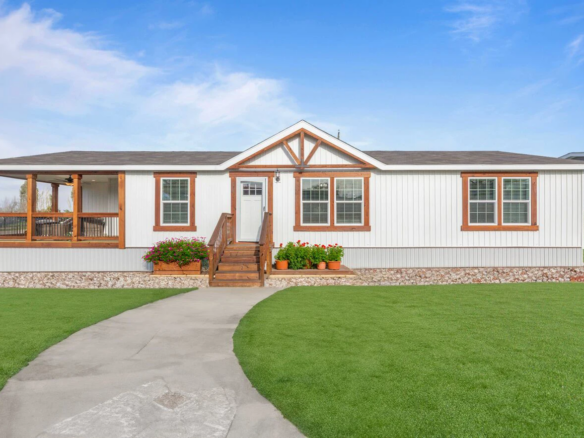 Single-story manufactured home with a front porch, white siding, brown trim, flower beds, and a concrete walkway across a well-kept lawn under a clear blue sky.