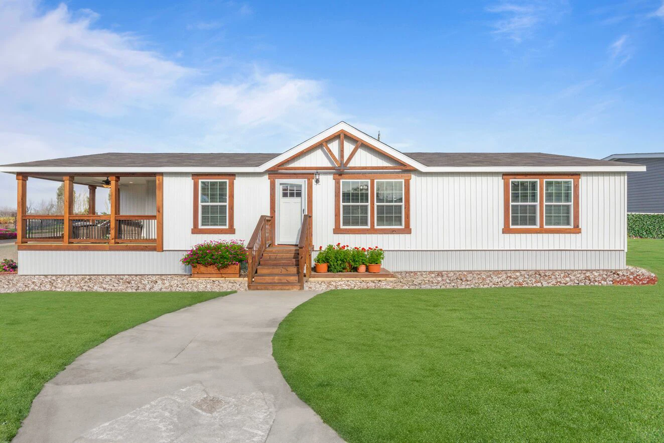 24cf551_1738879233573_1323_12 Single-story manufactured home with a front porch, white siding, brown trim, flower beds, and a concrete walkway across a well-kept lawn under a clear blue sky.