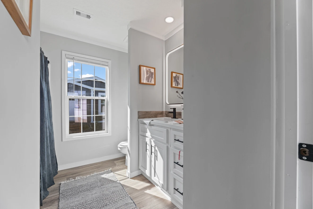 25_c_290_helix_1763n_bath_001_web_1742588944199_1323_12 A modern bathroom with a large window, white vanity with double sinks, framed mirror, two wall pictures, gray rug, and wood-look flooring.