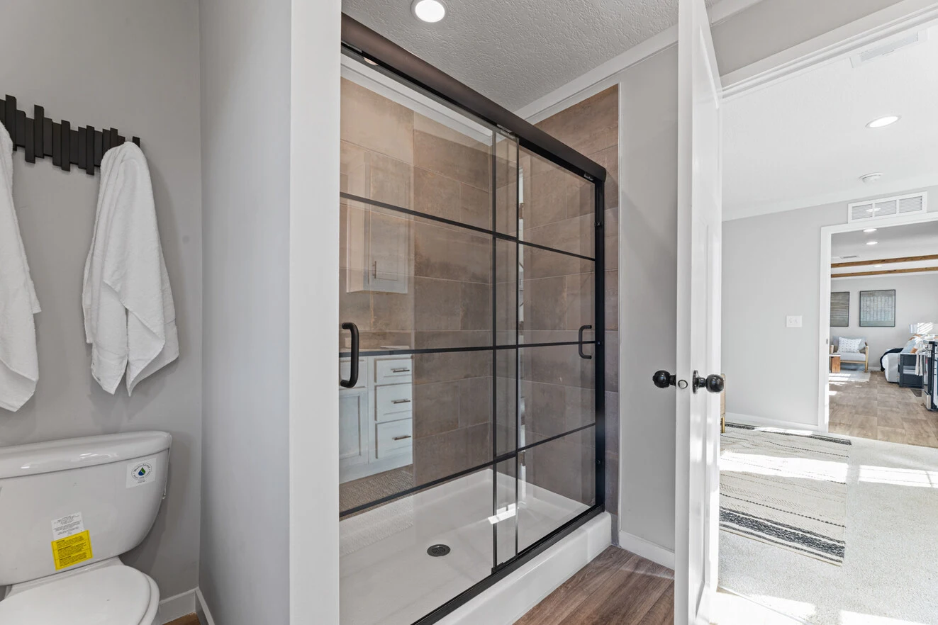 25_c_290_helix_1763n_bath_002_web_1742588941672_1323_12 Modern bathroom with a glass-enclosed shower featuring black trim, a white toilet, towel rack with two towels, and view into a bright adjoining room.