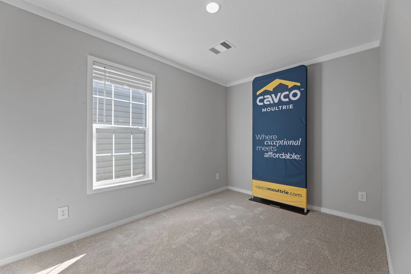 25_c_290_helix_1763n_bed_001_web_1742588932757_1323_12 Empty carpeted room with light gray walls, a window on the left, and a Cavco Moultrie promotional display standing against the right wall.