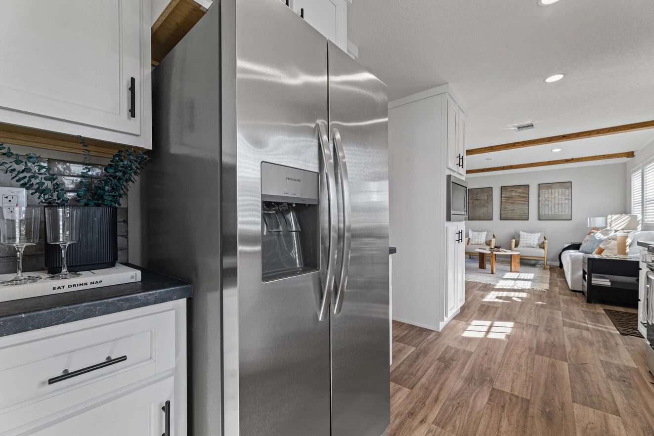 25_c_290_helix_1763n_kitchen_002_web_1742588911511_1323_12 Modern kitchen with a stainless steel refrigerator, white cabinets, wood flooring, and a view into a living room with sofas and wall art.