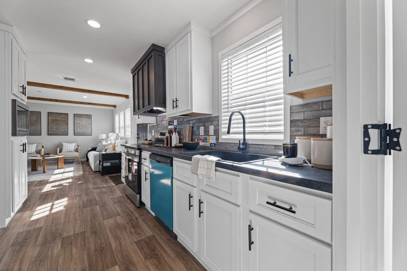 25_c_290_helix_1763n_kitchen_003_web_1742588909619_1323_12 Modern kitchen with white cabinets, black countertops, stainless steel appliances, blue dishwasher, and wood flooring, open to a living area with wall art and a sofa.