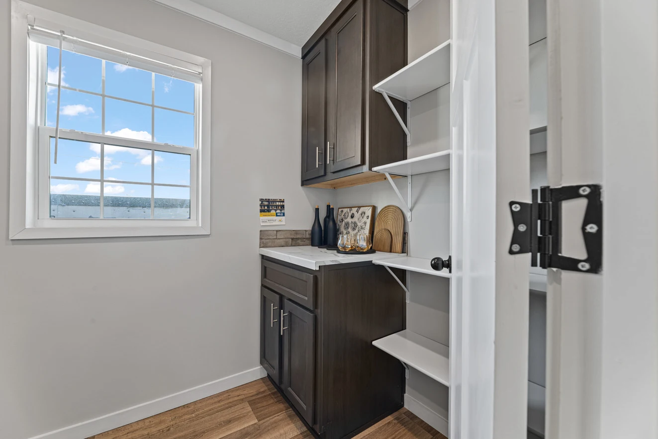 25_c_290_helix_1763n_utility_001_web_1742588989236_1323_12 Small pantry with open white shelves, dark wood cabinets, a countertop with bottles and cutting boards, and a window letting in natural light.
