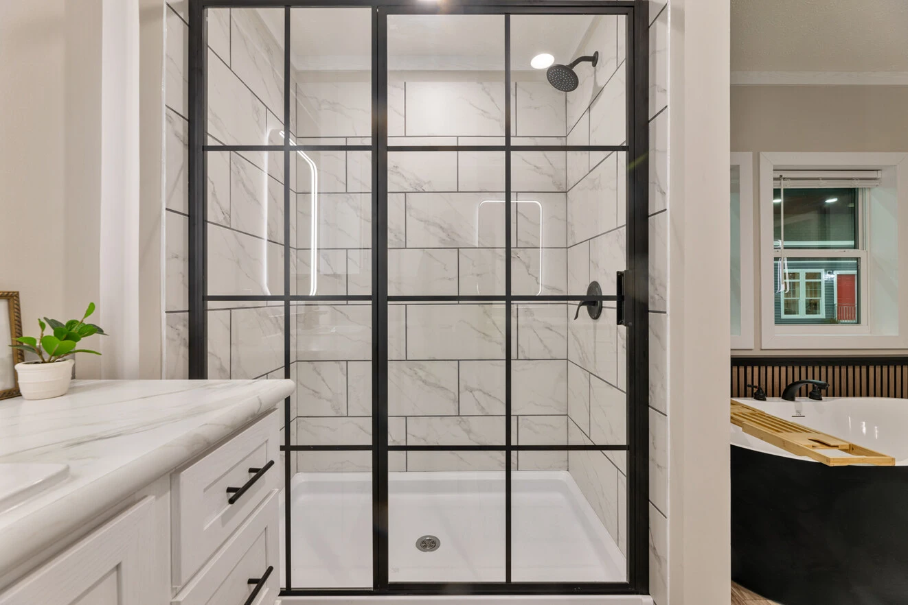 Glass shower enclosure with black grid frame, white marble tile interior, black shower fixtures, and part of a bathtub and sink visible nearby.