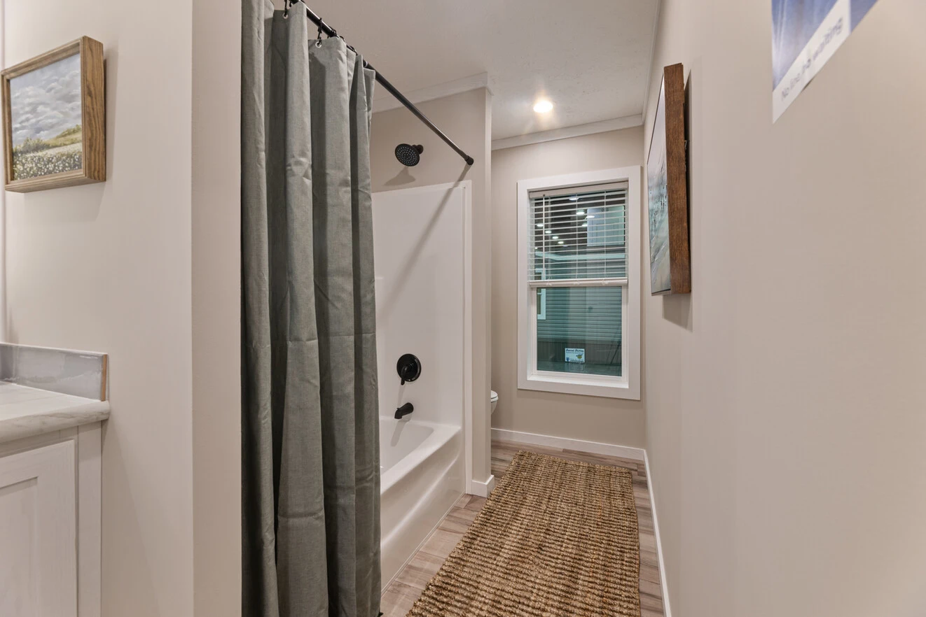 Narrow bathroom with a bathtub and shower, green curtain, white walls, framed art, a window, and a woven rug on the floor.