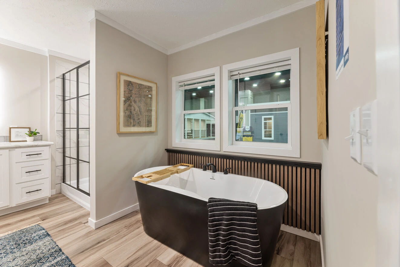 Modern bathroom with a black freestanding tub, wooden tray, striped towel, two windows, and light wood flooring next to a shower and white vanity.