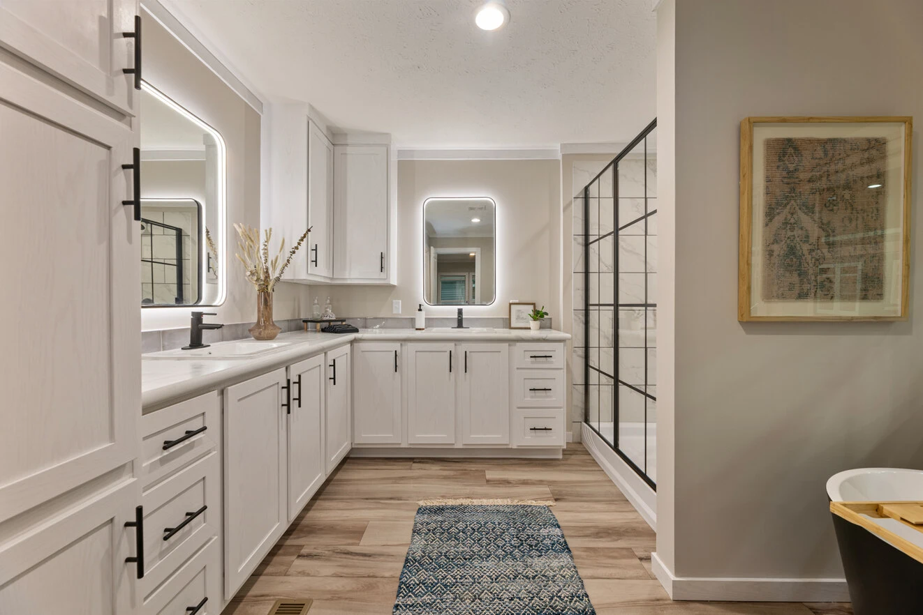 Modern bathroom with white cabinets, two large mirrors, a glass shower enclosure, wood-look flooring, and a blue patterned rug. A framed picture hangs on the right wall.