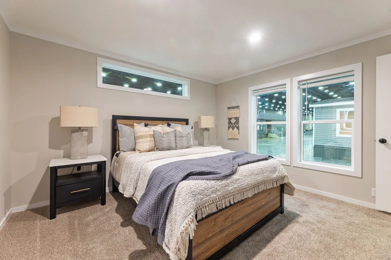 A modern bedroom with a neatly made bed, two nightstands with lamps, beige walls, carpeted floor, and large windows showing lights outside.
