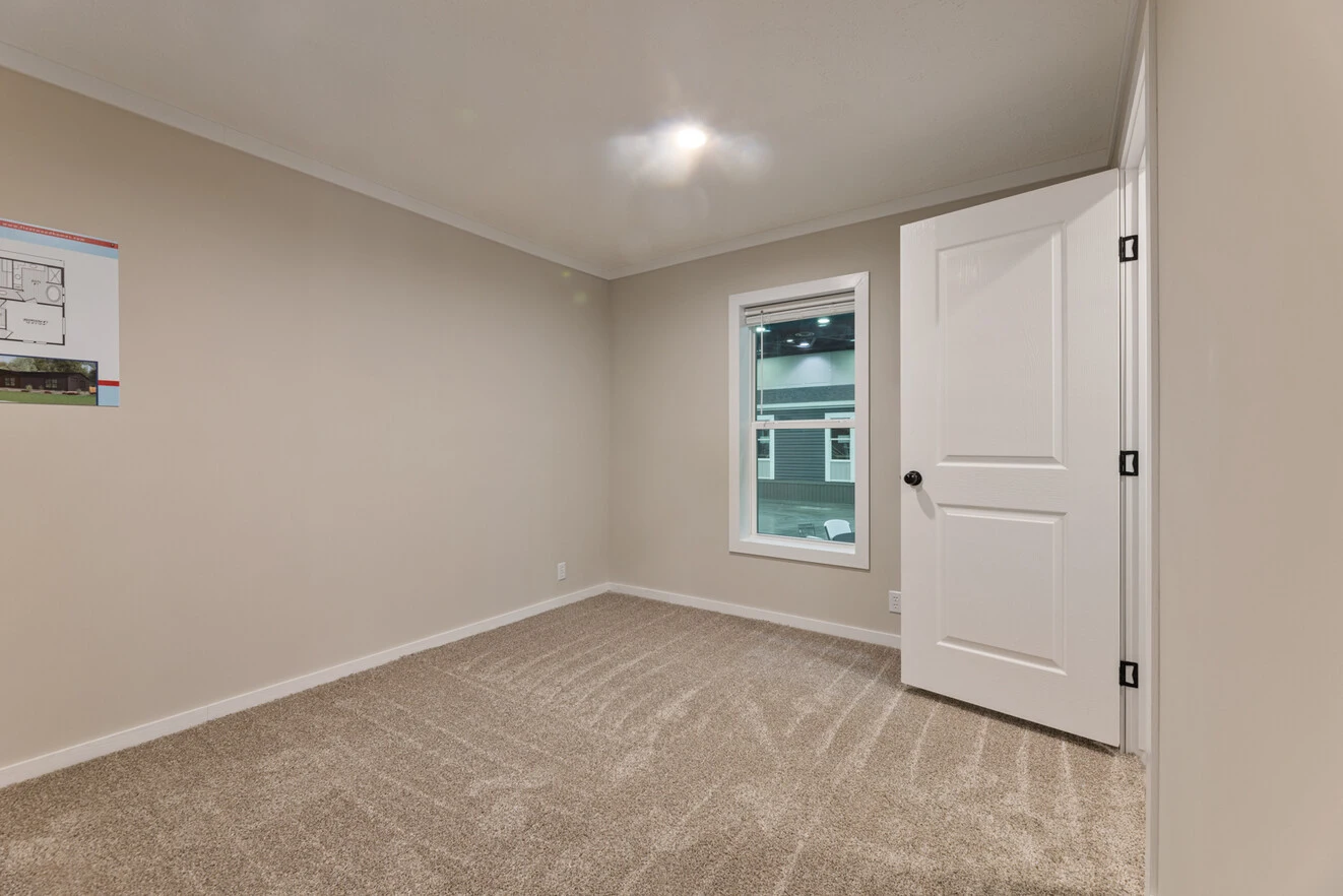 Empty carpeted room with beige walls, a closed white door, a window, and a wall-mounted floor plan diagram. Ceiling light is turned on.