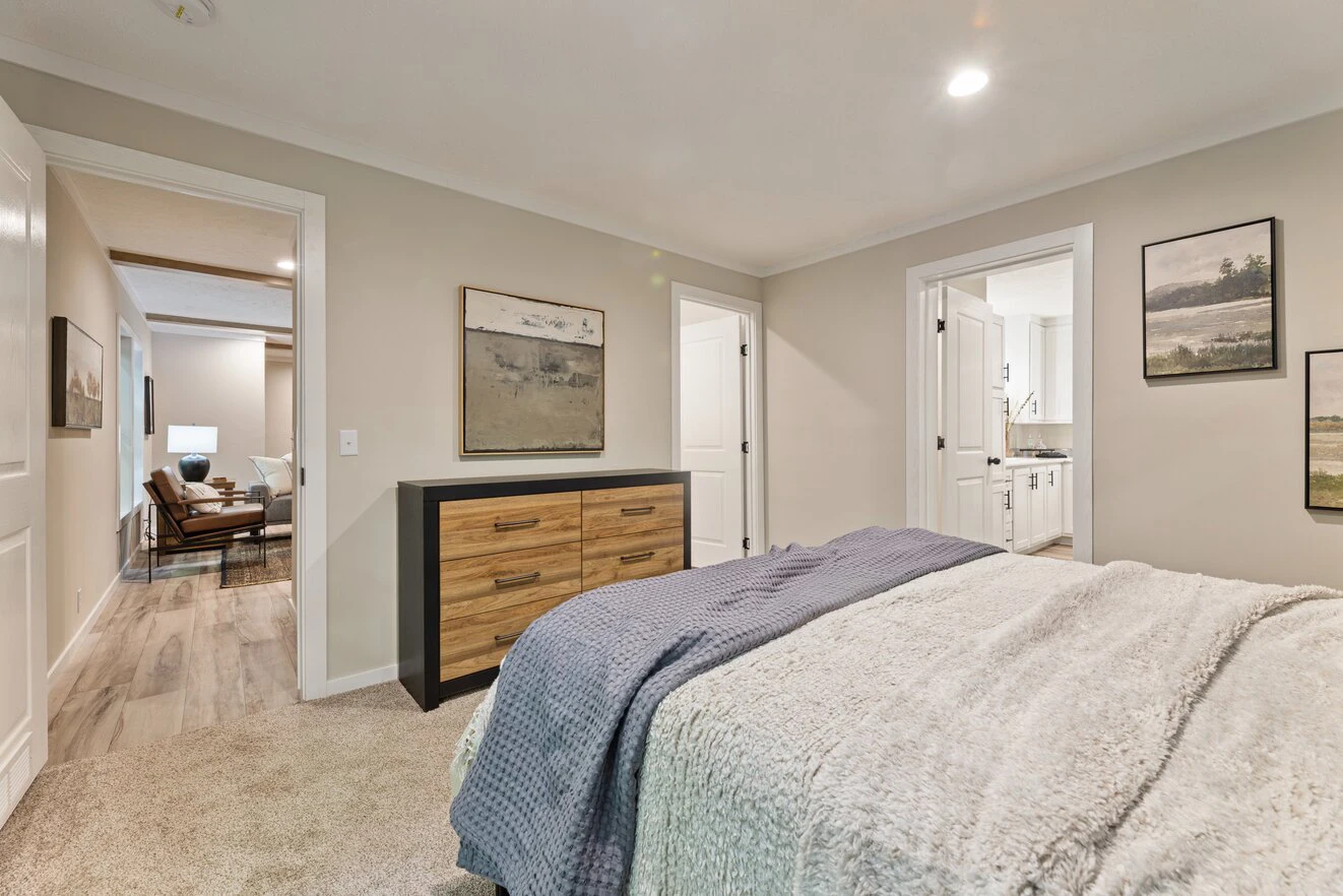 Modern bedroom with a neatly made bed, wooden dresser, neutral wall art, and open doors leading to a hallway and bathroom.
