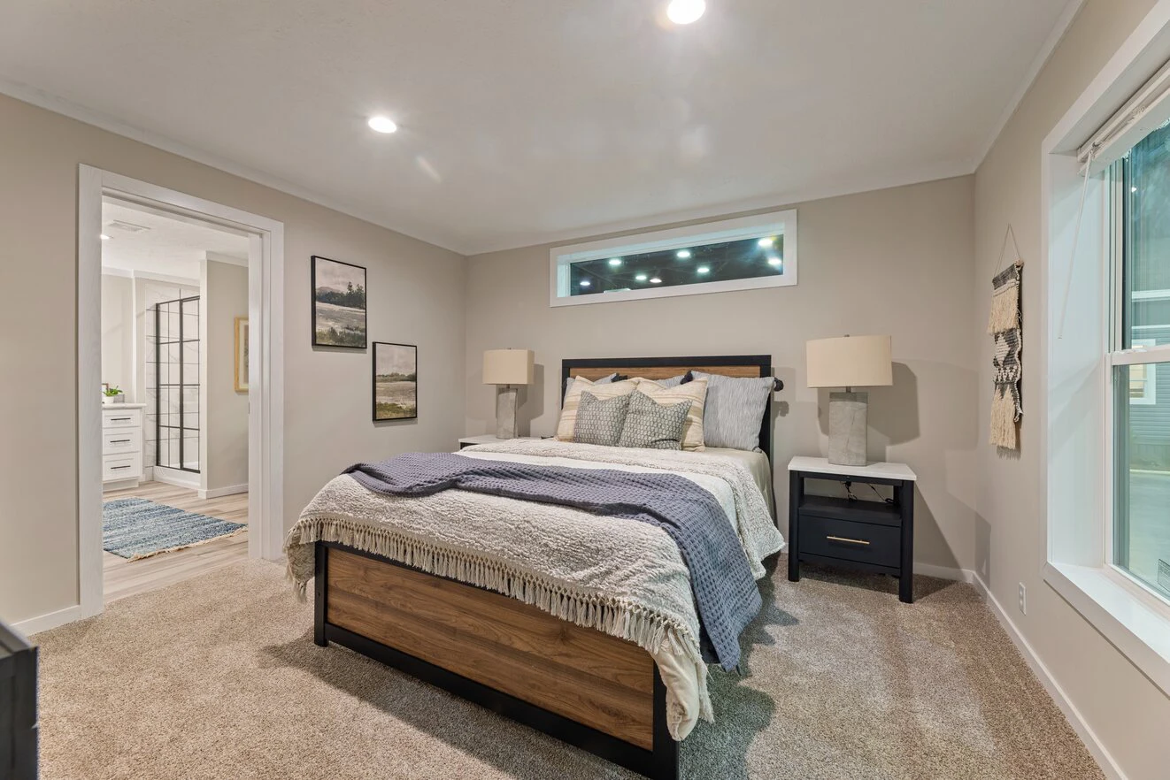 A modern bedroom with a bed, nightstands, lamps, wall art, and a large window, featuring neutral colors and carpeted flooring.