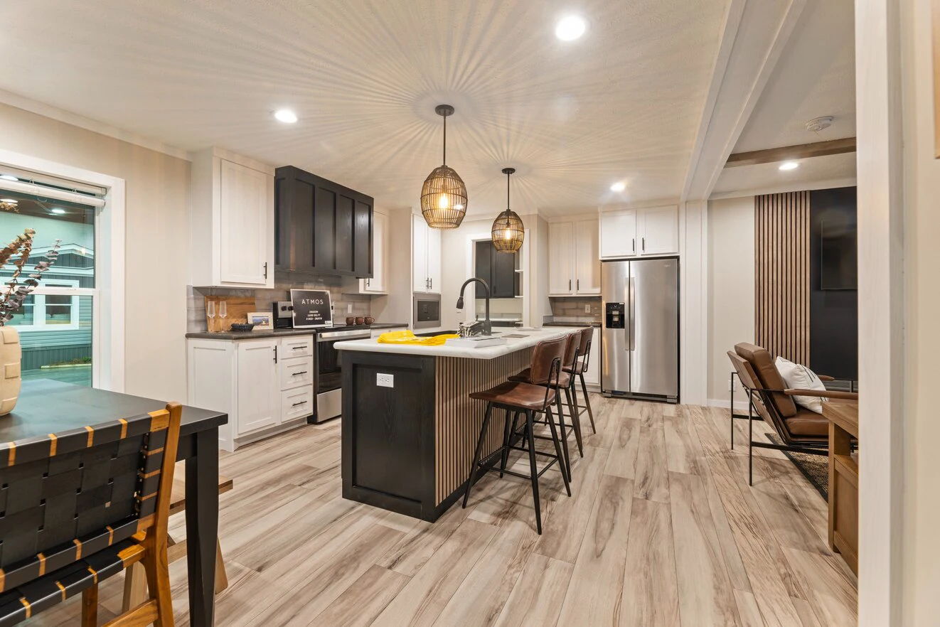 Modern kitchen with a central island, bar stools, stainless steel appliances, pendant lights, and a dining table to the left, featuring wood-style flooring and neutral decor.