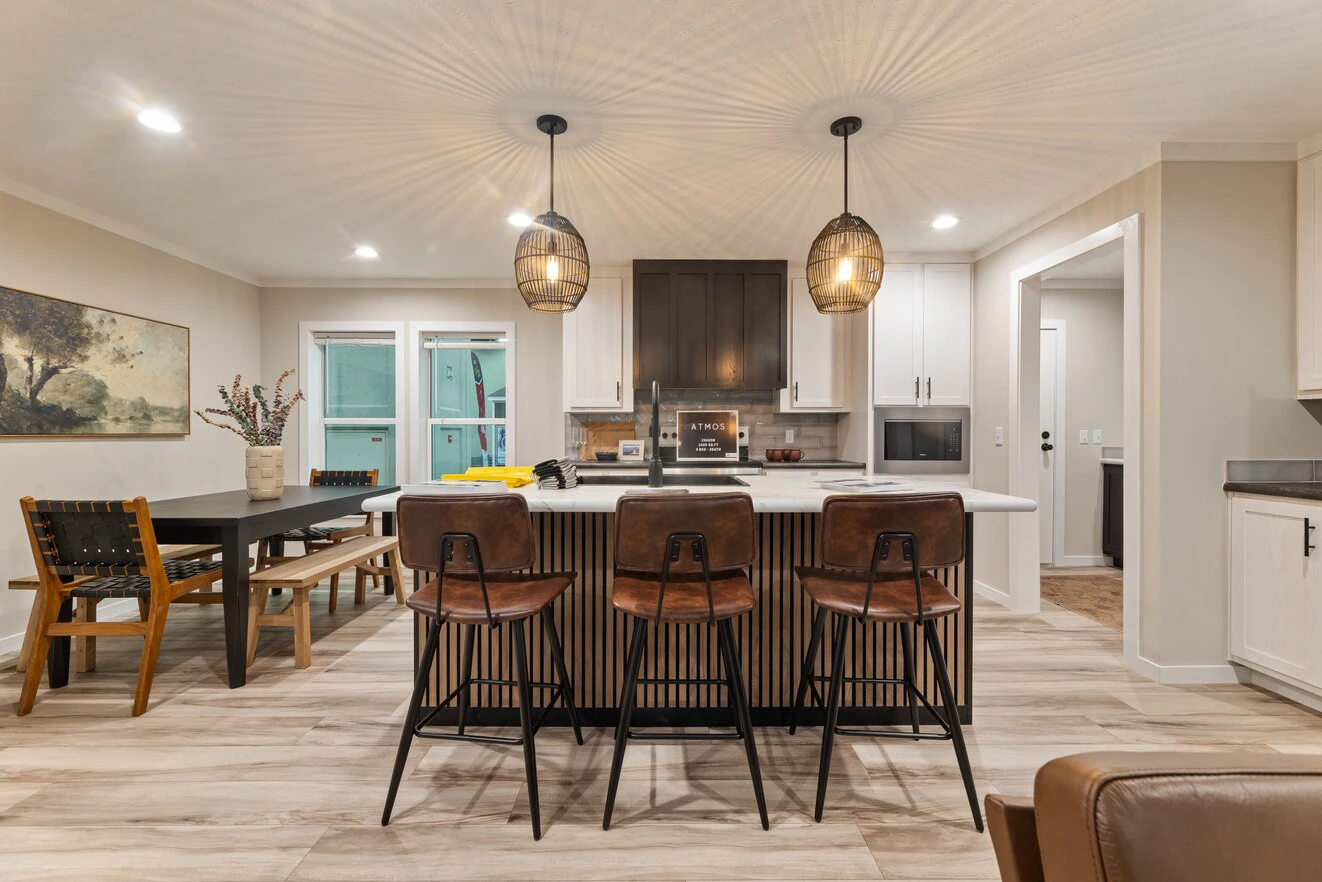 Modern kitchen with a central island, three brown barstools, pendant lights, a dining table with chairs, and neutral-colored cabinets and walls.