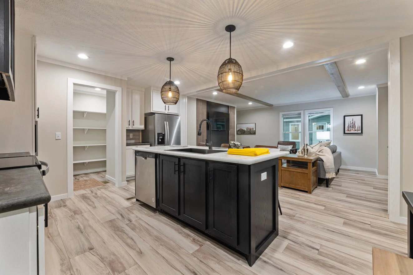 Modern kitchen with a central island, pendant lights, stainless steel appliances, open pantry, and a view into a furnished living room with wood-style flooring.