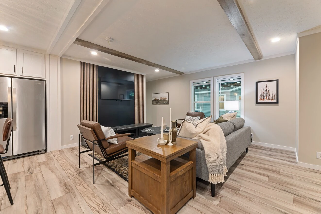 Modern living room with wood accents, gray sofa, black chairs, a large TV, and a side table with candles, decorated in neutral tones with natural light from two windows.