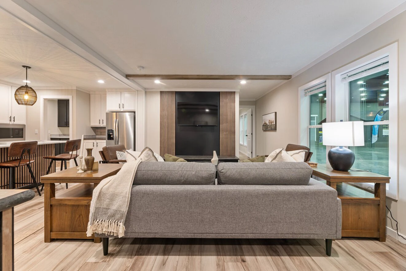 Modern living room with a gray sofa, wooden furniture, flat-screen TV, and open kitchen in the background, featuring neutral colors and large windows.