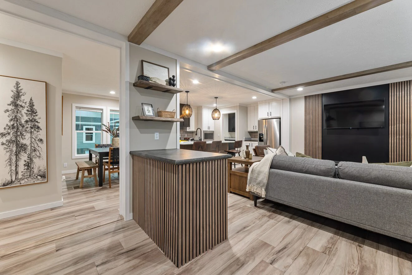 Open-concept living space with wood accents, modern kitchen with pendant lights, dining area, gray sectional sofa, and wall-mounted TV. Light wood flooring throughout.