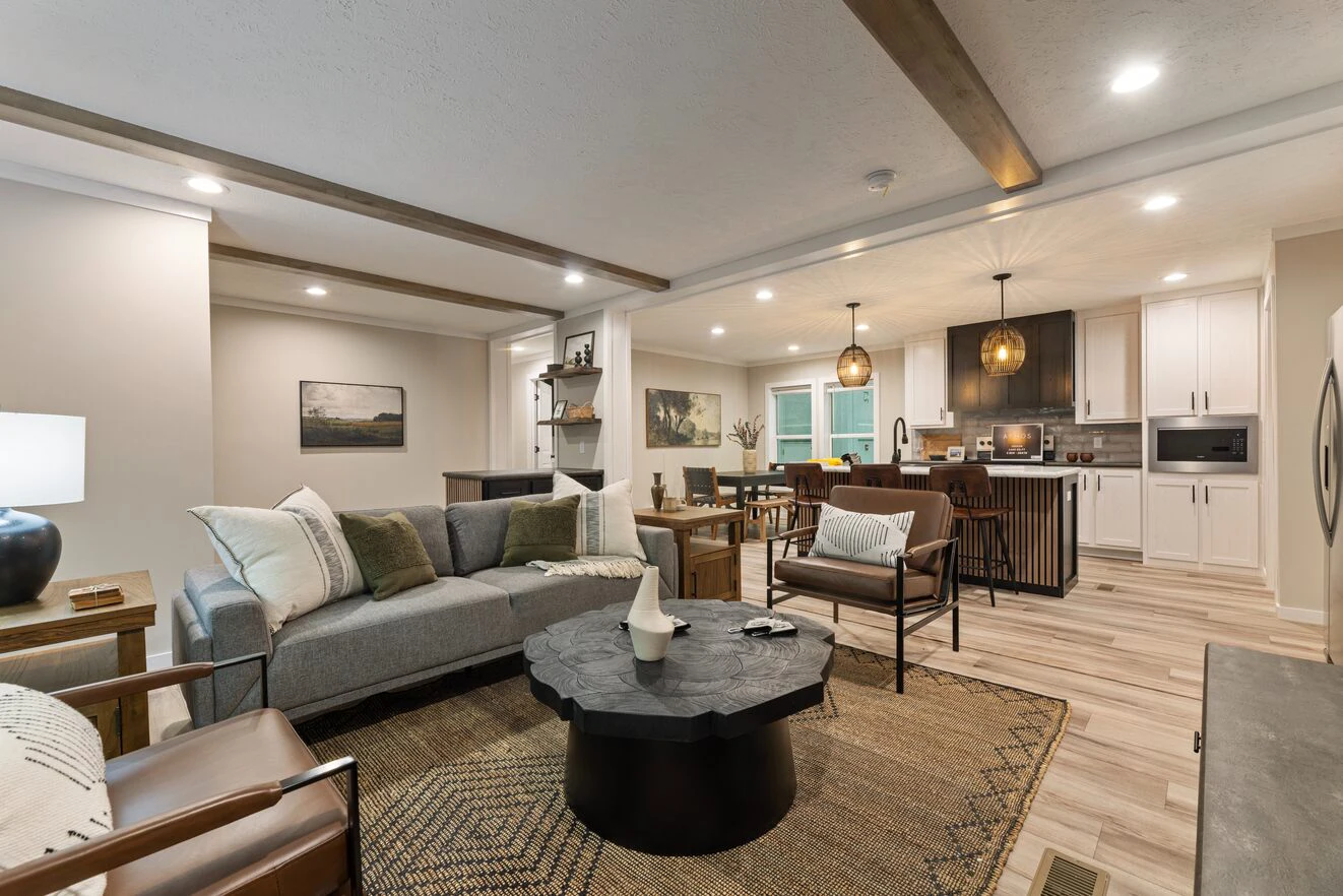 Modern open-concept living room and kitchen with neutral tones, wooden accents, a gray sofa, round black coffee table, and pendant lights over the island.