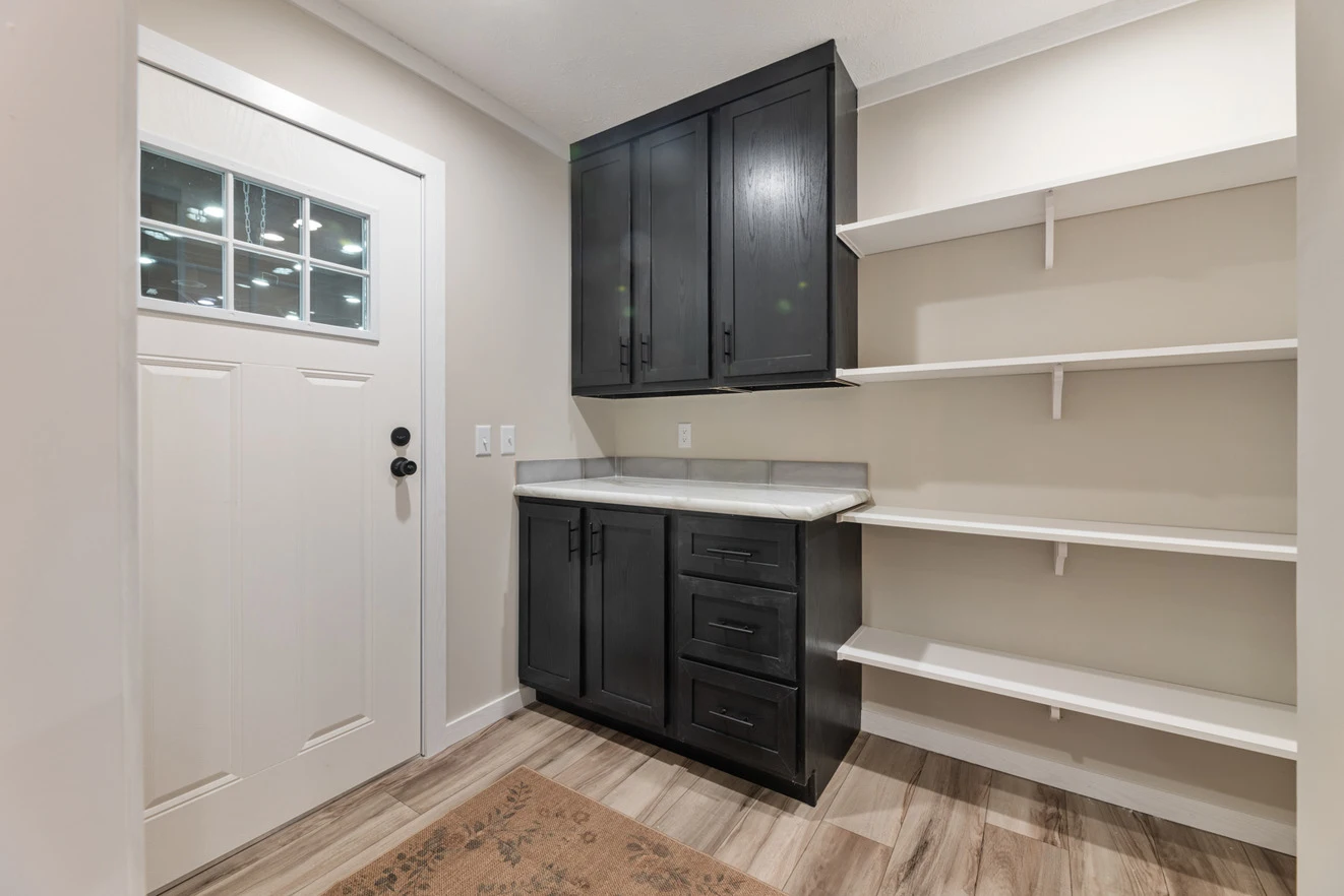 A small room with a white door, black cabinets with a marble countertop, and white wall-mounted shelves on wood-patterned flooring.