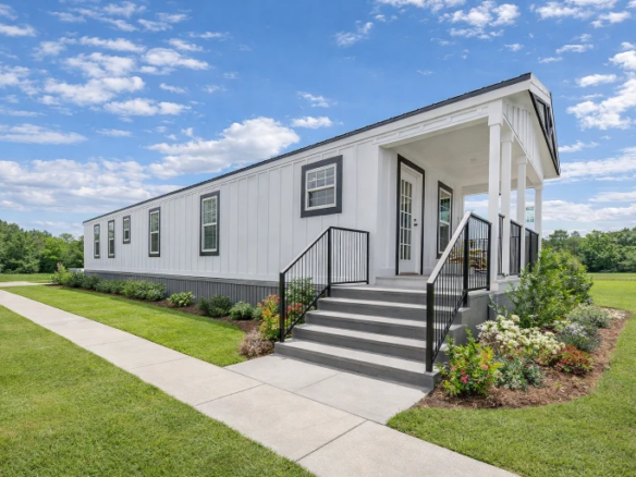 A modern single-story manufactured home with white siding, a front porch, black railings, and landscaped yard under a partly cloudy sky.