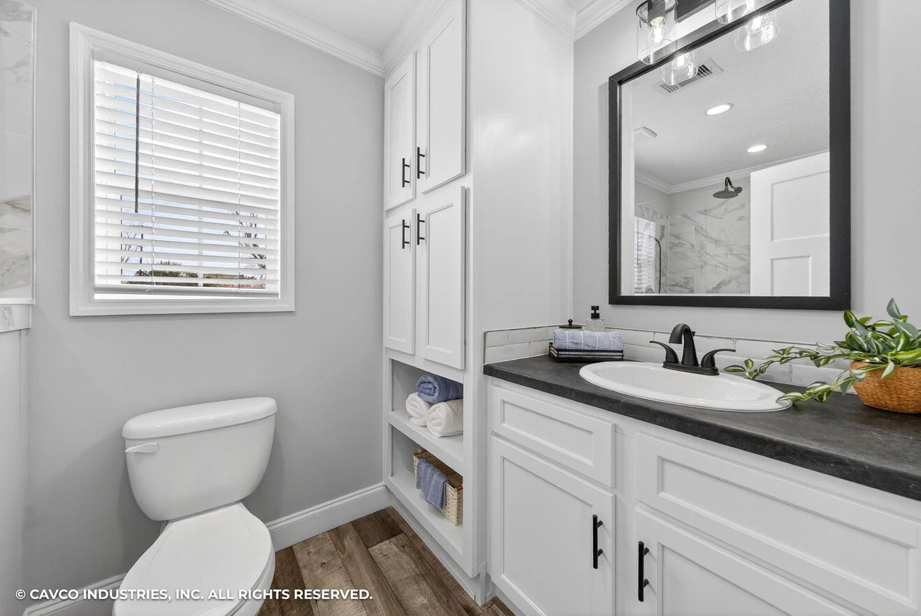 Modern bathroom with a white toilet, built-in storage, a sink with a black faucet, a potted plant, and a window with blinds. The floor is wood-style and walls are light gray.