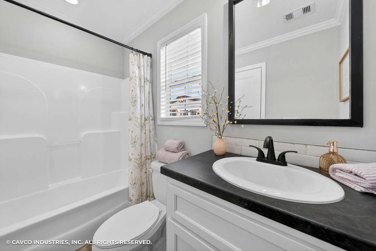 Modern bathroom with a white bathtub and shower, black countertop with sink, mirror, folded towels, and a vase with branches on the vanity.