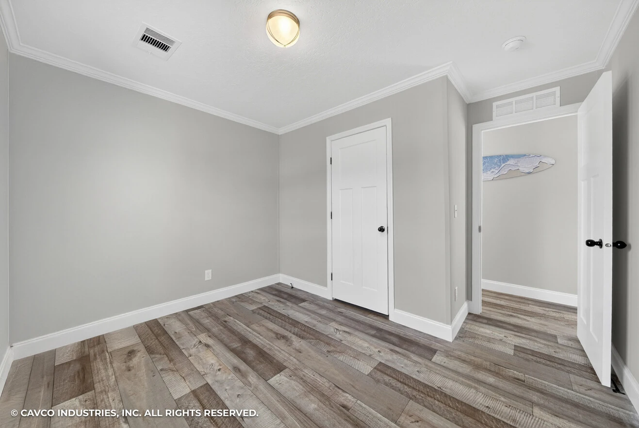 Empty room with light gray walls, white trim, wood-style flooring, a ceiling light, and two closed white doors; a hallway with a wall art piece is visible through one door.