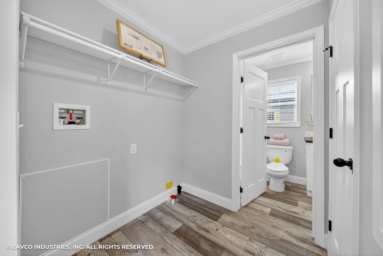 Laundry room with wall shelf, washer and dryer hookups, and a doorway leading to a small bathroom with a toilet and window.