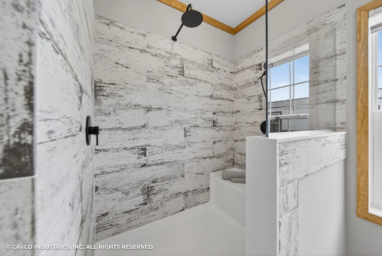 26_c_290_satilla_ly32764d_bath_002_web_copyright_1774469462053_1320_12 Modern bathroom shower with a rainfall showerhead, textured white and gray tile walls, built-in bench, and a window letting in natural light.