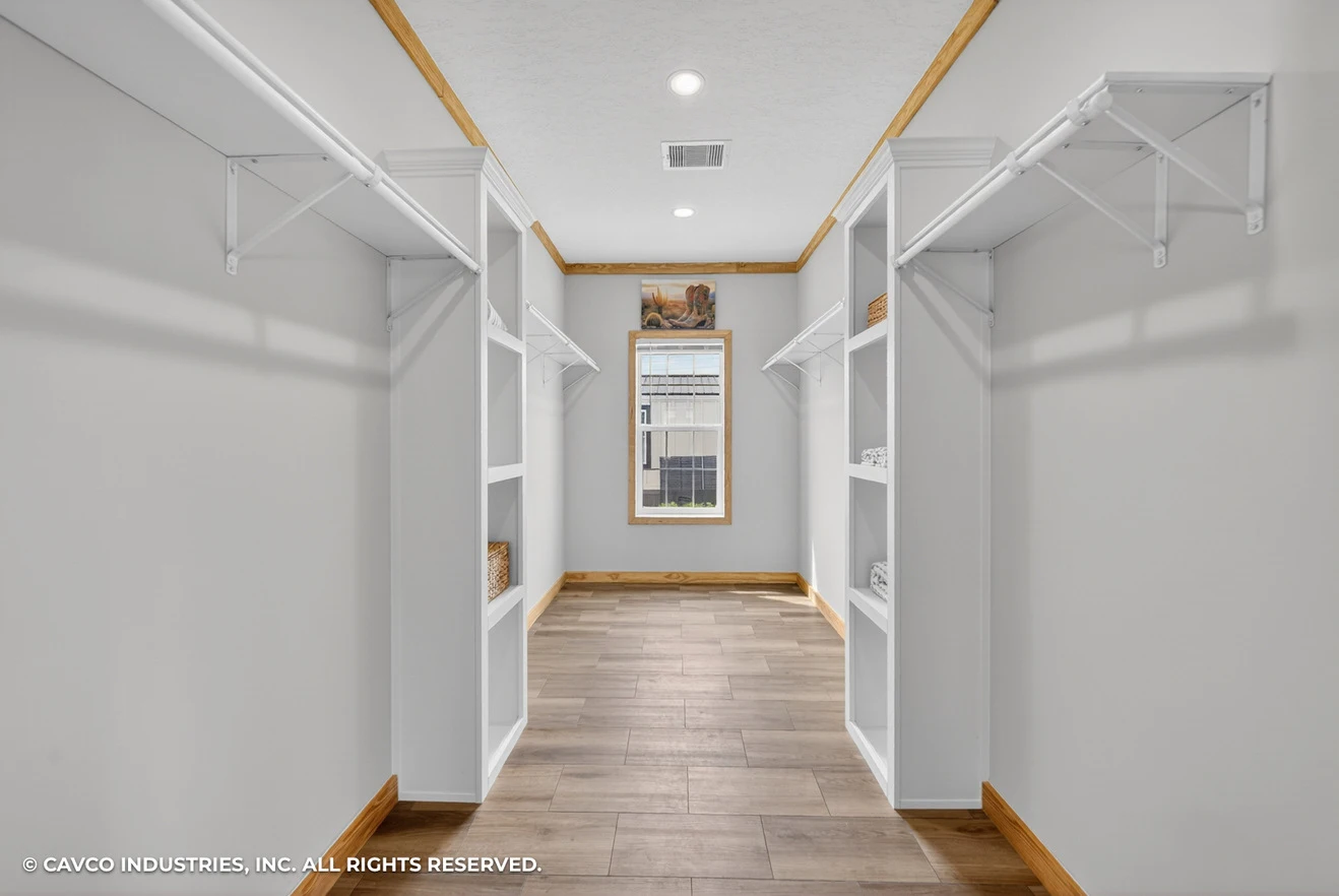 26_c_290_satilla_ly32764d_bed_007_web_copyright_1774469429884_1319_12 Spacious walk-in closet with built-in white shelves, hanging rods, wood trim, and a central window letting in natural light.