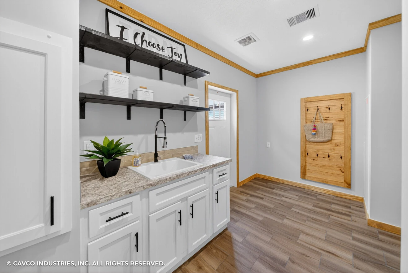 26_c_290_satilla_ly32764d_kitchen_001_web_copyright_1774469417449_1317_12 Laundry room with white cabinets, a farmhouse sink, open black shelves, and a wooden accent door with a basket hanging on it. Marble countertop and wood-style flooring.