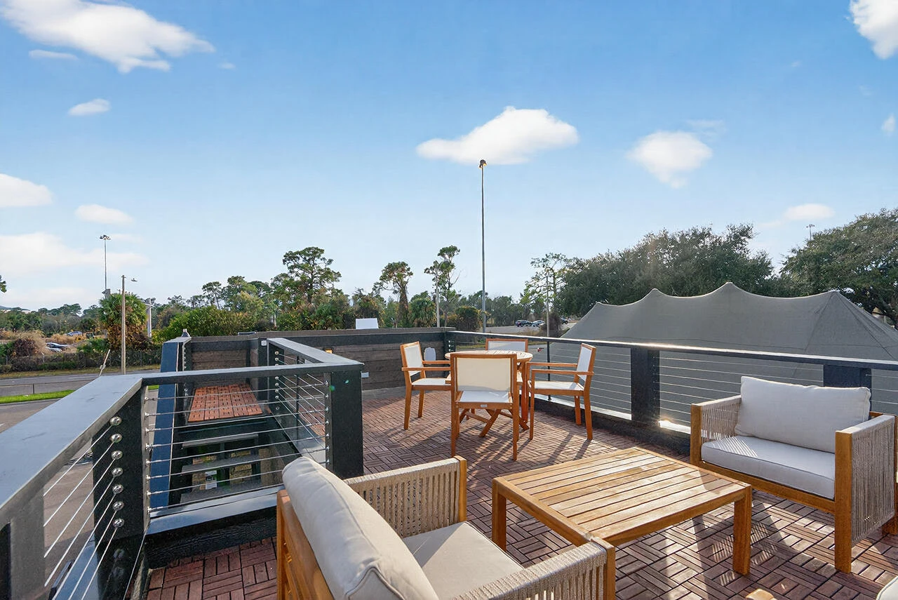 Outdoor rooftop patio with wooden furniture, including chairs and a table, surrounded by a railing, under a blue sky with scattered clouds.
