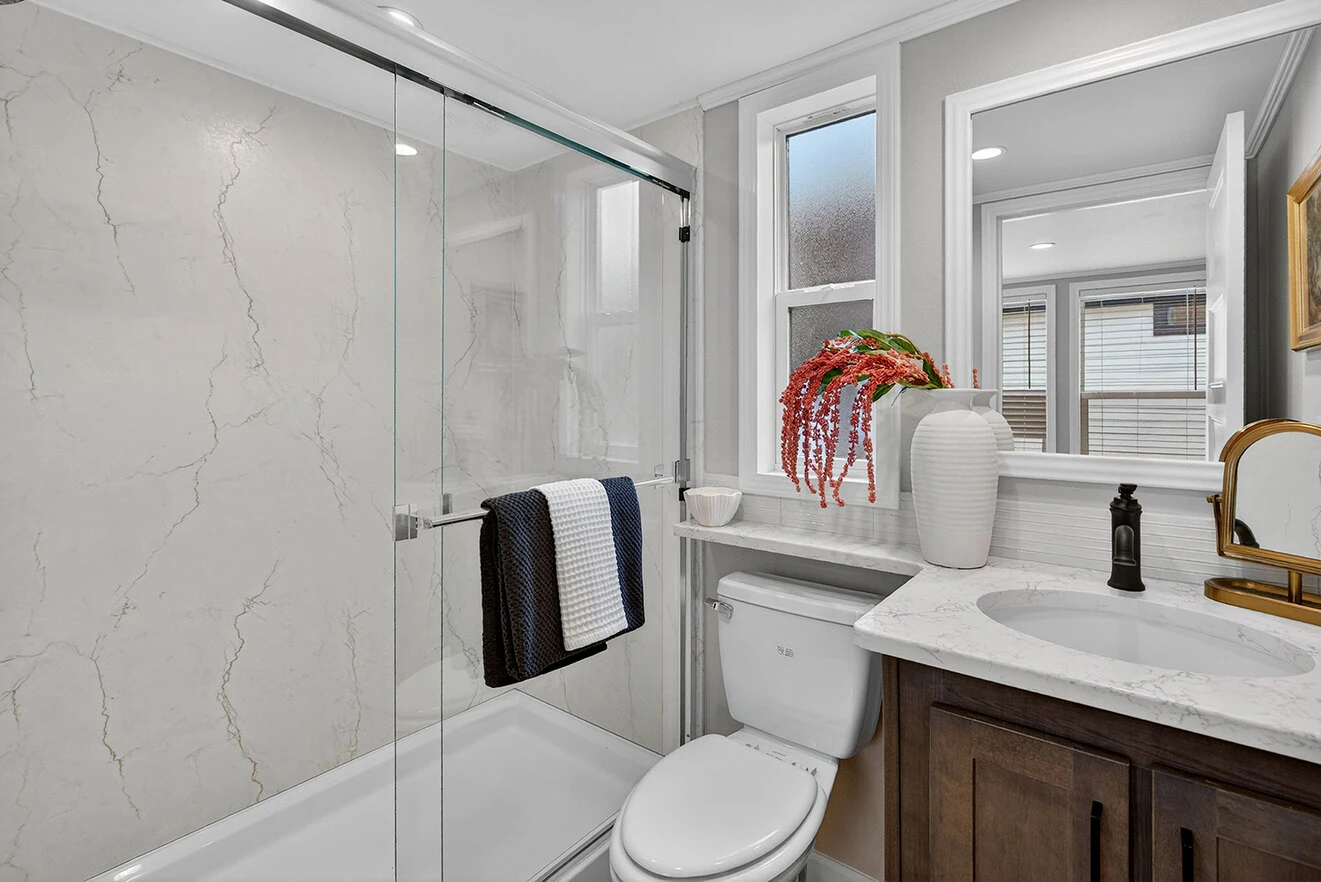Modern bathroom with a glass-enclosed shower, toilet, vanity with sink, gold faucet, and white countertop; decorative towels and a vase with red flowers on the counter.