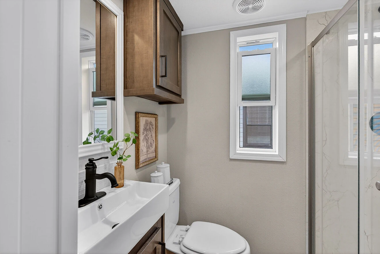 Small modern bathroom with a white sink, black faucet, toilet, wall cabinet, glass shower, and two narrow windows letting in natural light.