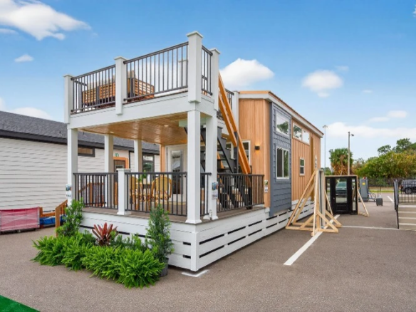 A modern tiny house with wood paneling features a covered porch and rooftop deck, surrounded by plants and parking spaces, under a blue sky.