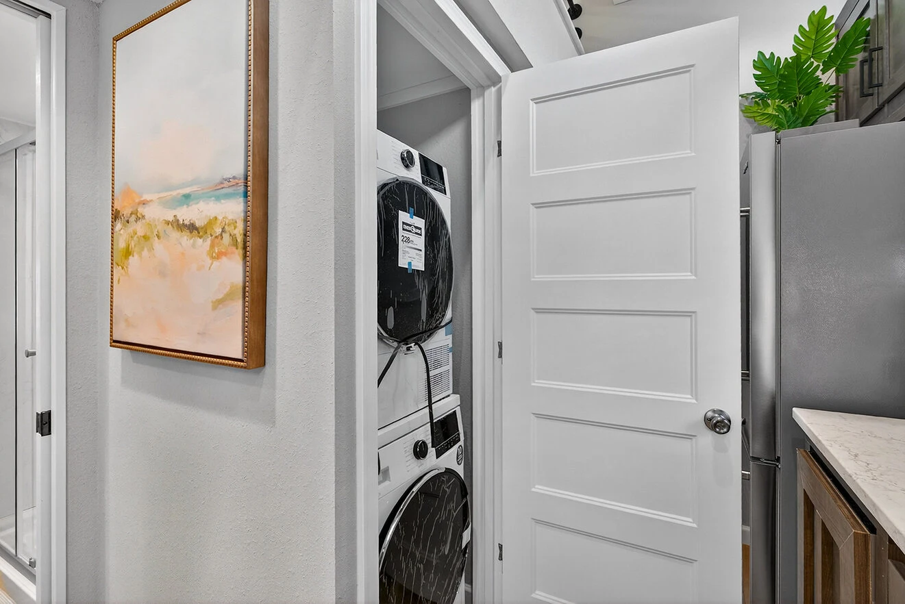 A white door opens to reveal a stacked washer and dryer in a small laundry closet next to a hallway with a framed landscape painting on the wall.