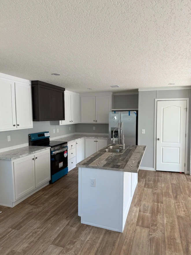 28684a_iron_ranch_2_1756133574303_661_12 Modern kitchen with white cabinets, a gray island, stainless steel refrigerator, and a blue-covered stove. The floor is wooden, and the walls are light gray with a white door.
