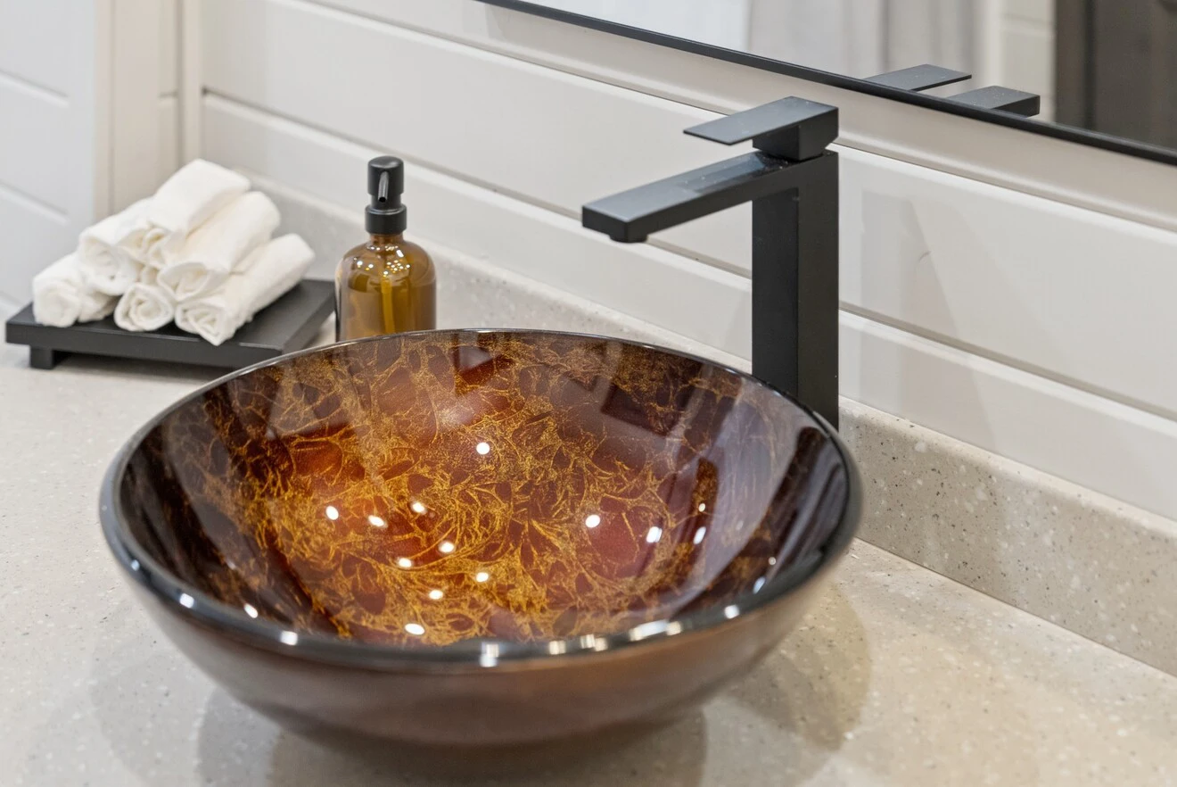 Modern bathroom countertop with a round glass vessel sink in amber tones, a black faucet, a soap dispenser, and rolled white towels next to a mirror.