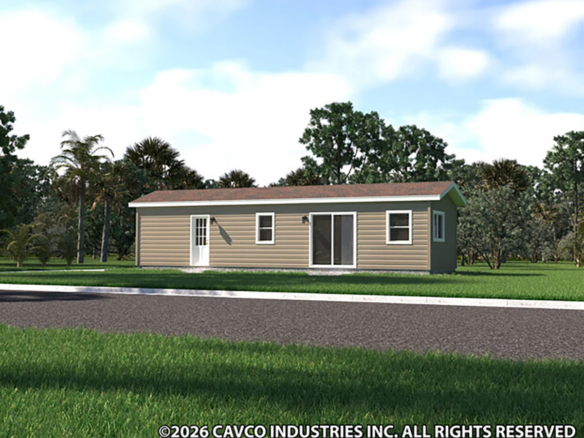A small, single-story manufactured home with tan siding and white trim sits on a grassy lot surrounded by trees.