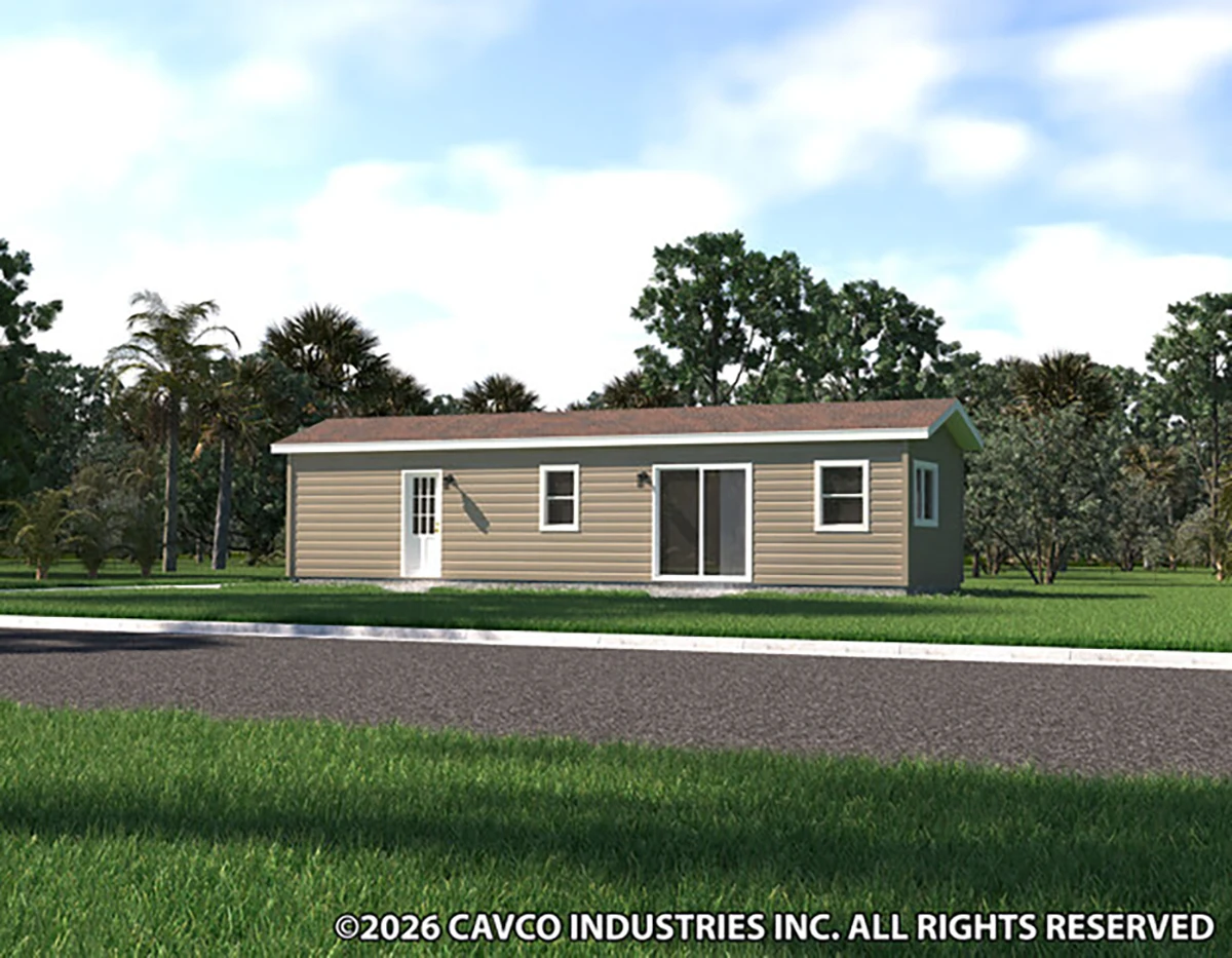 A small, single-story manufactured home with tan siding and white trim sits on a grassy lot surrounded by trees.