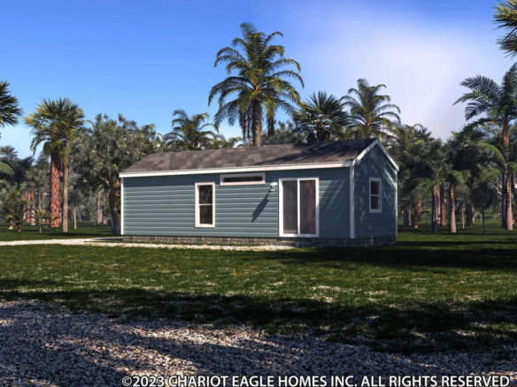A small, single-story blue manufactured home surrounded by grass, palm trees, and a partly cloudy sky overhead.