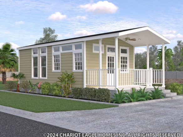A small, single-story modular home with beige siding, large windows, a covered porch, and surrounding plants under a partly cloudy sky.