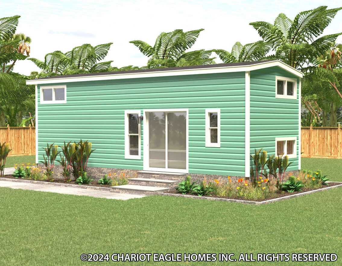 A small green modern tiny house with horizontal siding, large sliding glass doors, and plants in front, set in a fenced yard with tropical trees.