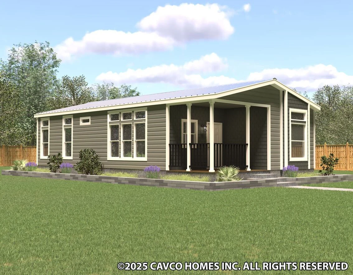 Modern single-story manufactured home with a covered front porch, large windows, and landscaped yard, set against a backdrop of trees and a wooden fence.
