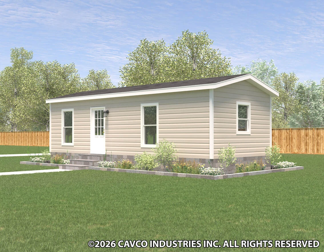 Small beige manufactured home with white trim, a gable roof, and a white door, set on a grassy lawn with flower beds and trees in the background.